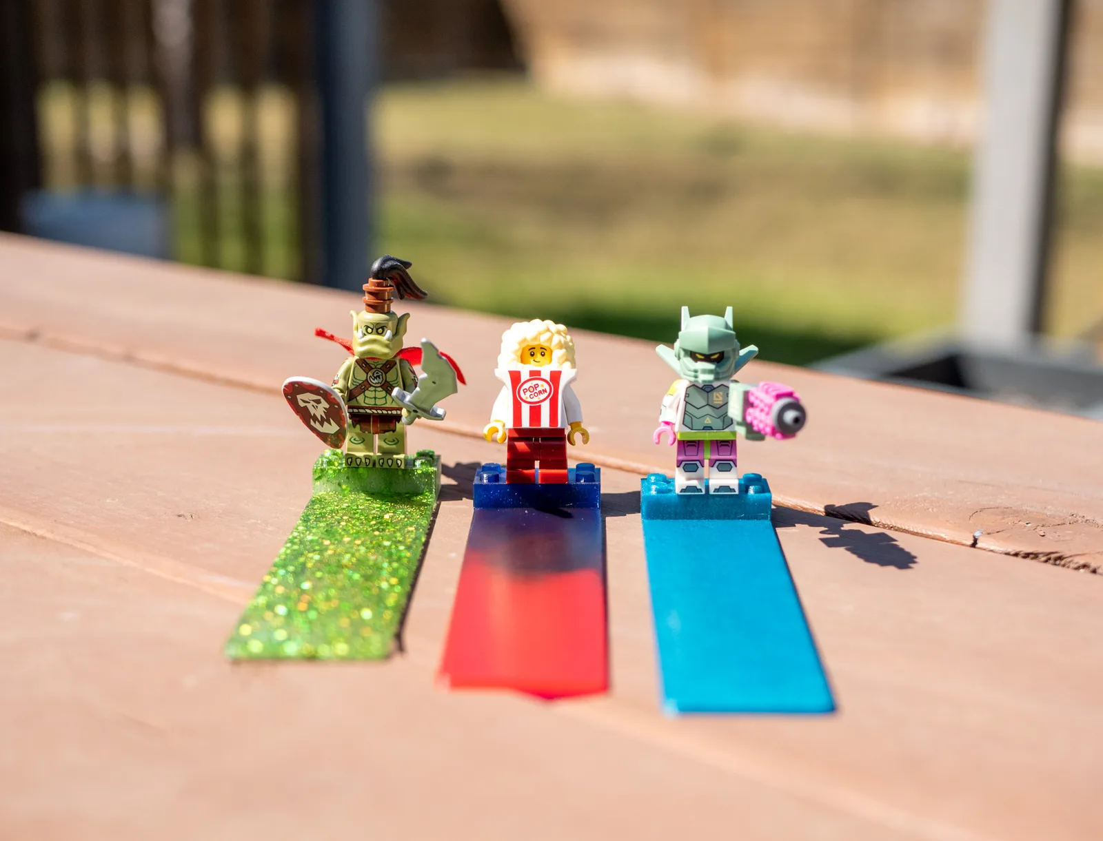 Three minifigure bookmarks in green, red and blue lined up on a wooden table outdoors
