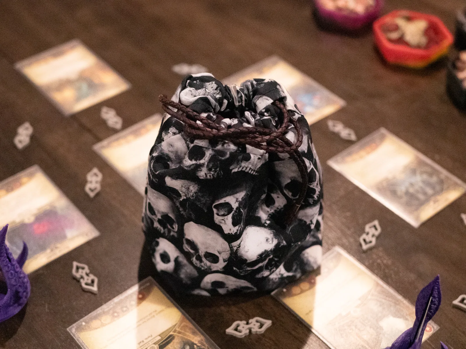 A skull-patterned drawstring bag standing on a game board surrounded by cards and tokens